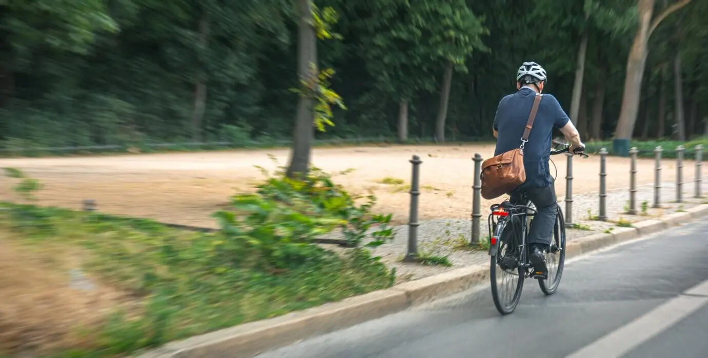 Radfahrer mit Tasche fährt am Park vorbei, nachhaltige Stadt, Bewegung.